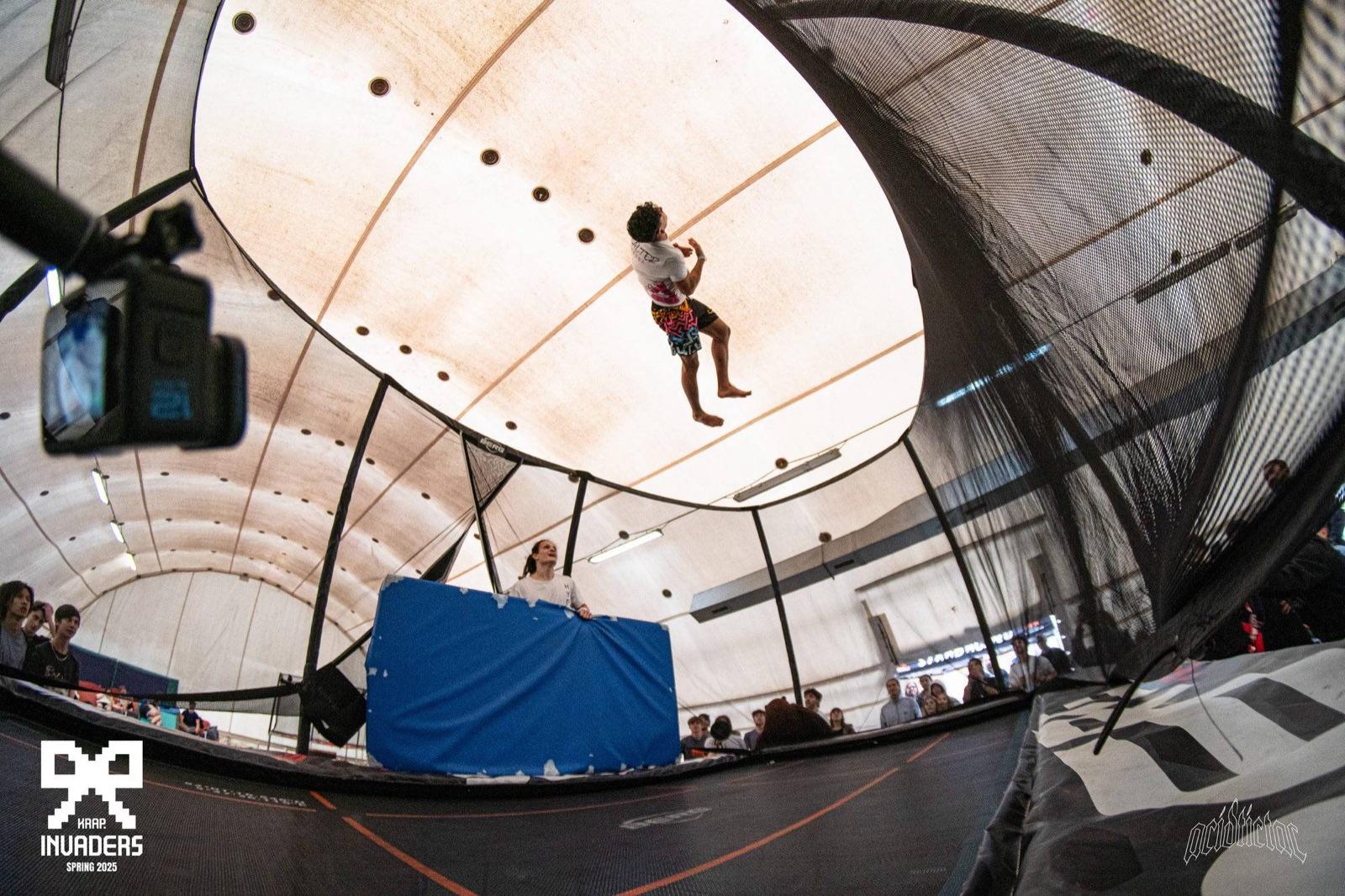 Un ragazzo in volo altissimo sul trampolino con la rete, sotto la tensostruttura, con il pubblico intorno